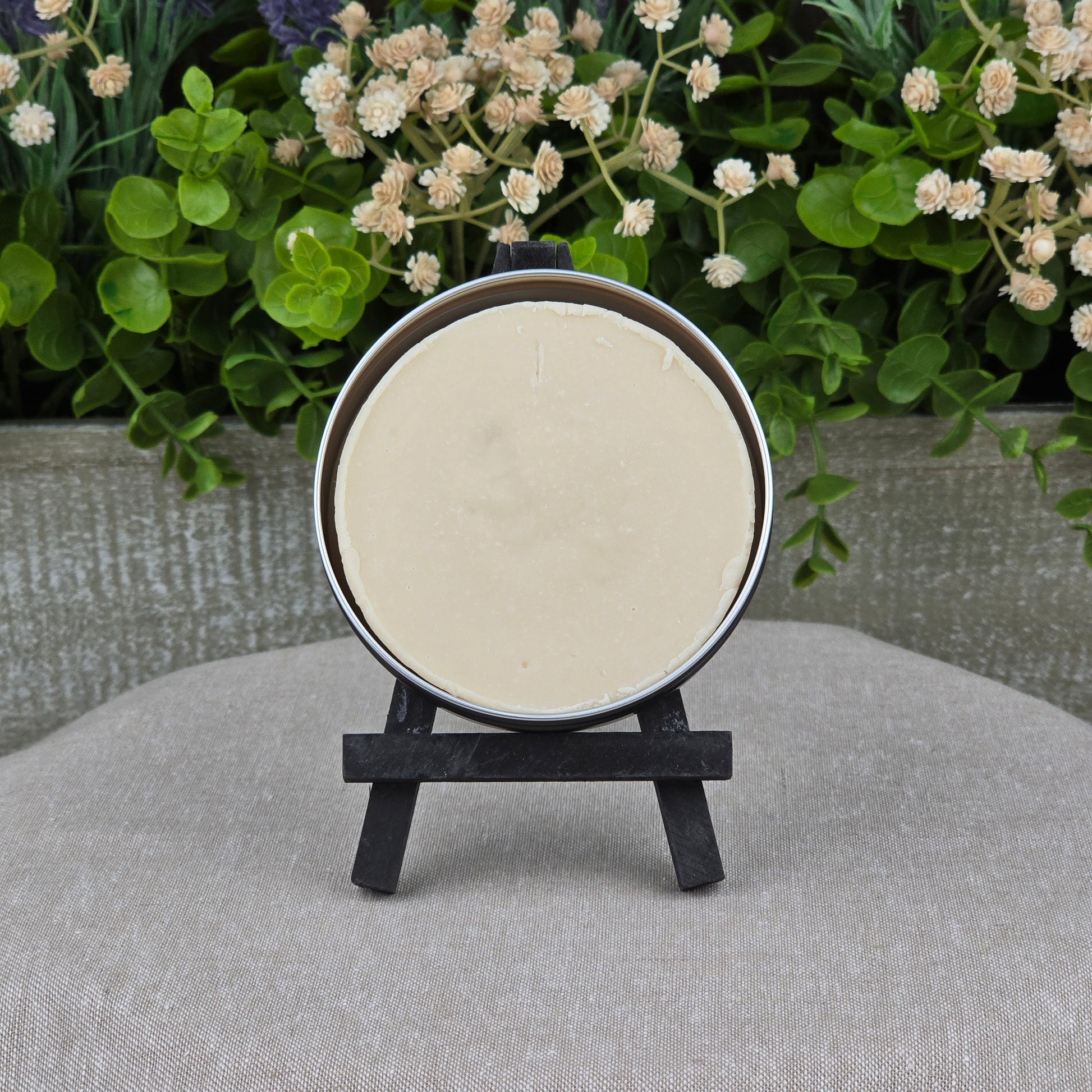 A round tin of Jarhead Aromatics Lavender Handmade Shave Soap with beef tallow and goat milk is displayed on a black easel among green leaves and white flowers atop beige fabric, perfect for wet shaving enthusiasts.
