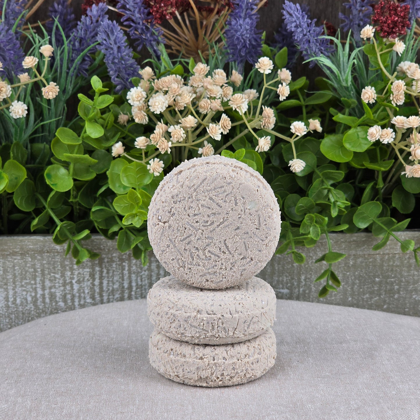 Three round Lavender Handmade Shampoo & Conditioner Bars by Jarhead Aromatics are stacked on a gray surface, surrounded by green leaves and purple and cream flowers—a beautiful, zero-waste hair care display.