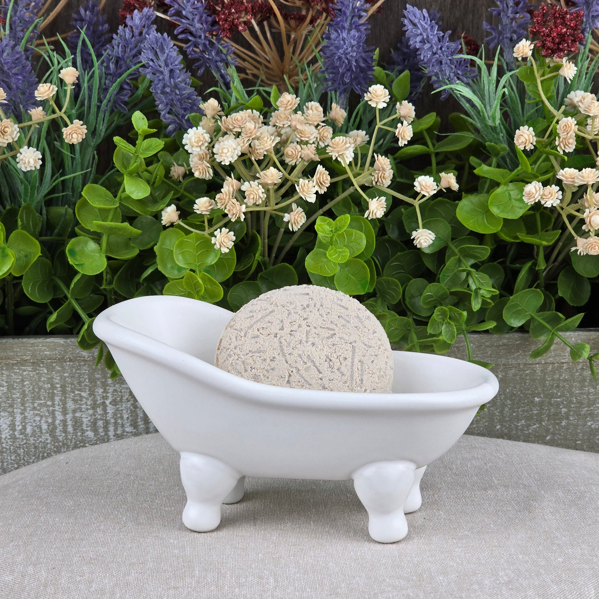 The Lavender Handmade Shampoo & Conditioner Bar by Jarhead Aromatics sits in a white ceramic tub on beige, surrounded by green plants and purple flowers—a tranquil scene ideal for zero-waste hair care enthusiasts.