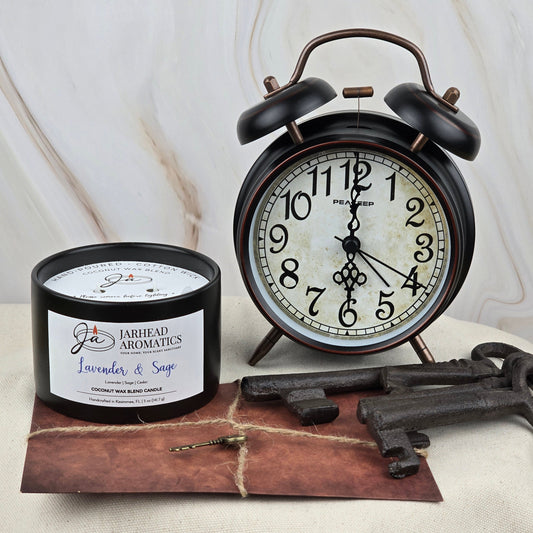 A black vintage alarm clock shows 6:00 beside the Jarhead Aromatics Timeless Collection Lavender & Sage Coconut Wax Blend Candle, evoking a subtle spa feel, with an old brown envelope and three antique keys nearby.