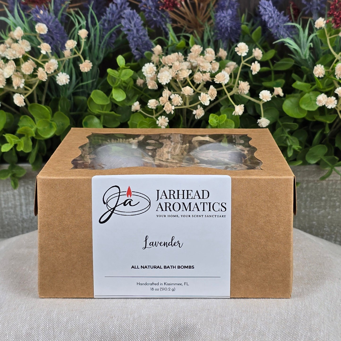 A brown box labeled Jarhead Aromatics Patchouli Bath Bomb - All Natural with cruelty-free bath bombs sits on a gray surface, a clear window revealing the bath bombs inside. Purple, white, and red artificial flowers and greenery are in the background.