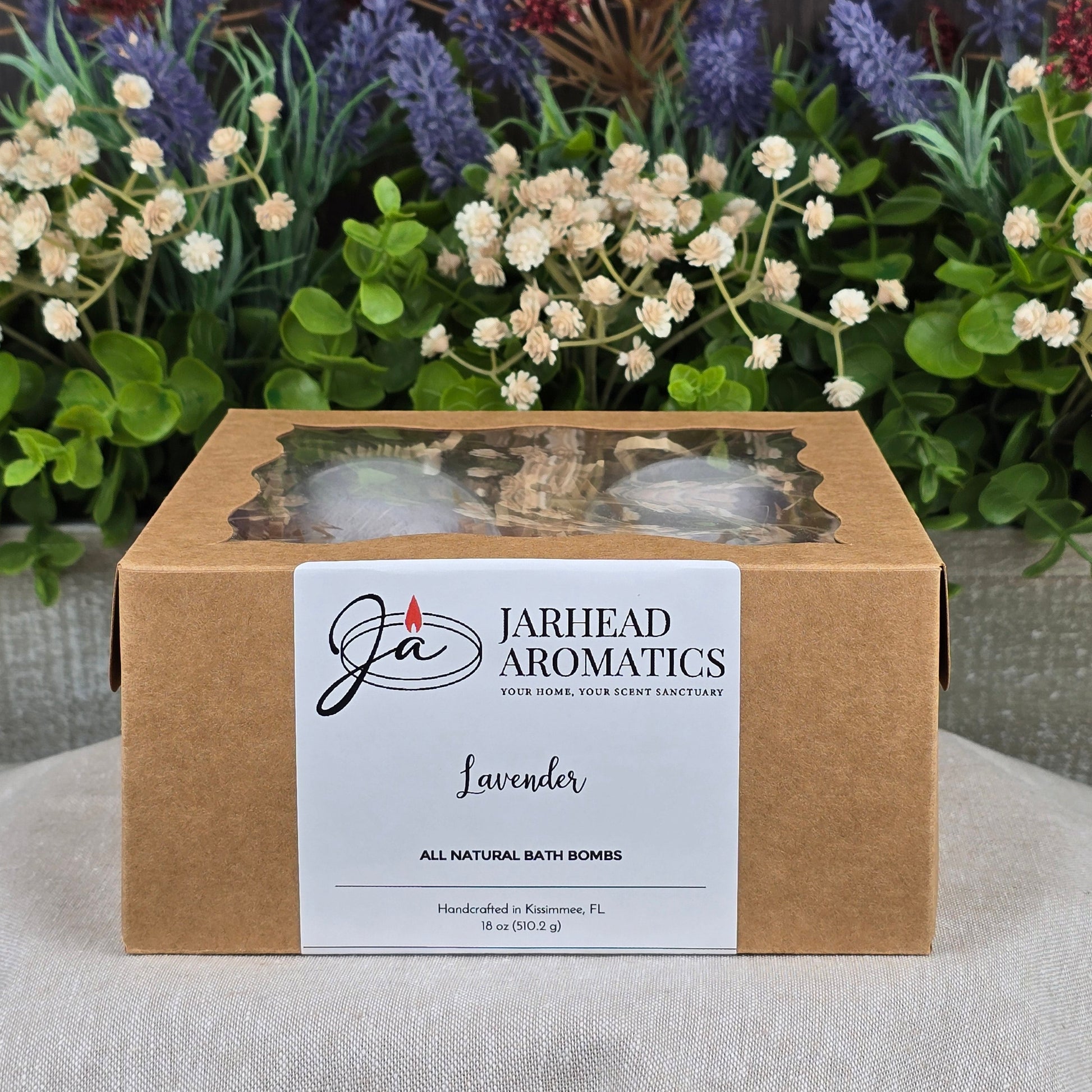 A brown box labeled Jarhead Aromatics Patchouli Bath Bomb - All Natural with cruelty-free bath bombs sits on a gray surface, a clear window revealing the bath bombs inside. Purple, white, and red artificial flowers and greenery are in the background.