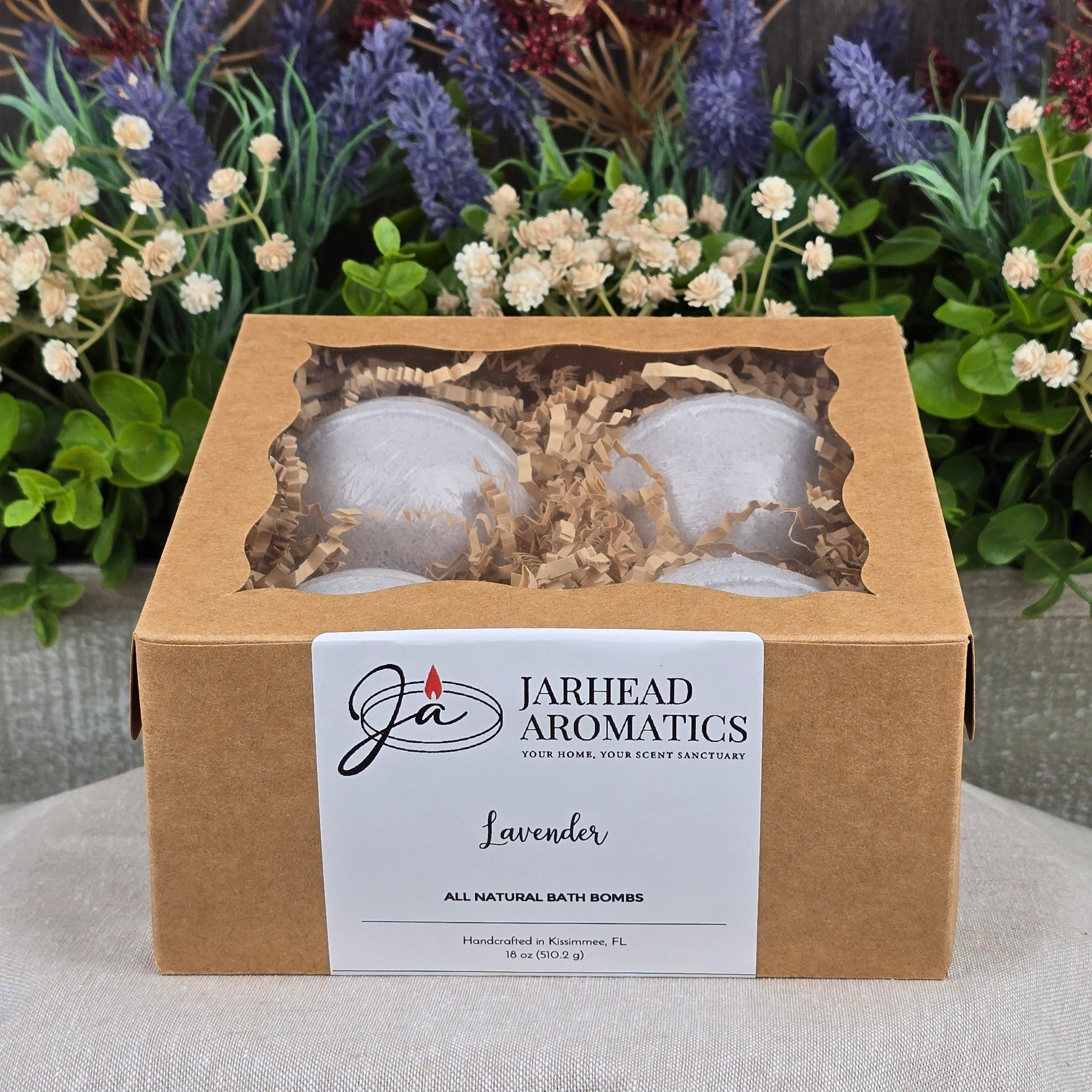 A box of Jarhead Aromatics Patchouli Bath Bomb - All Natural sits on a gray surface, set against purple and white flowers. The brown box has a clear window displaying cruelty-free bath bombs and a white label on the front.