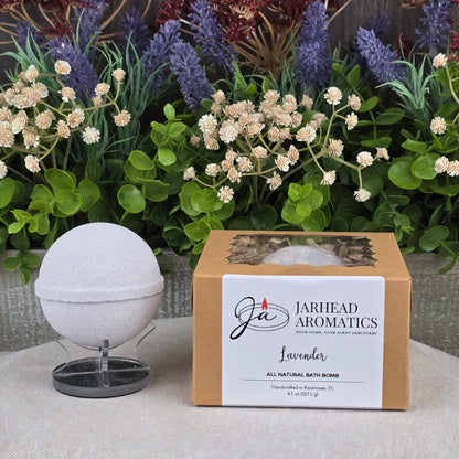 A white natural bath bomb on a stand sits next to a brown box labeled Jarhead Aromatics Patchouli Bath Bomb - All Natural (Copy), with assorted artificial flowers and greenery in the background.
