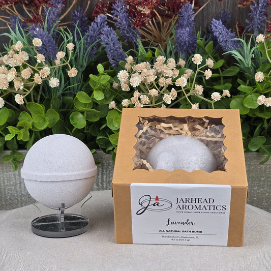 A handmade Patchouli Bath Bomb - All Natural from Jarhead Aromatics rests on a stand beside its branded box, which features a cutout displaying another bath bomb. The scene is accented with greenery and purple flowers in the background.