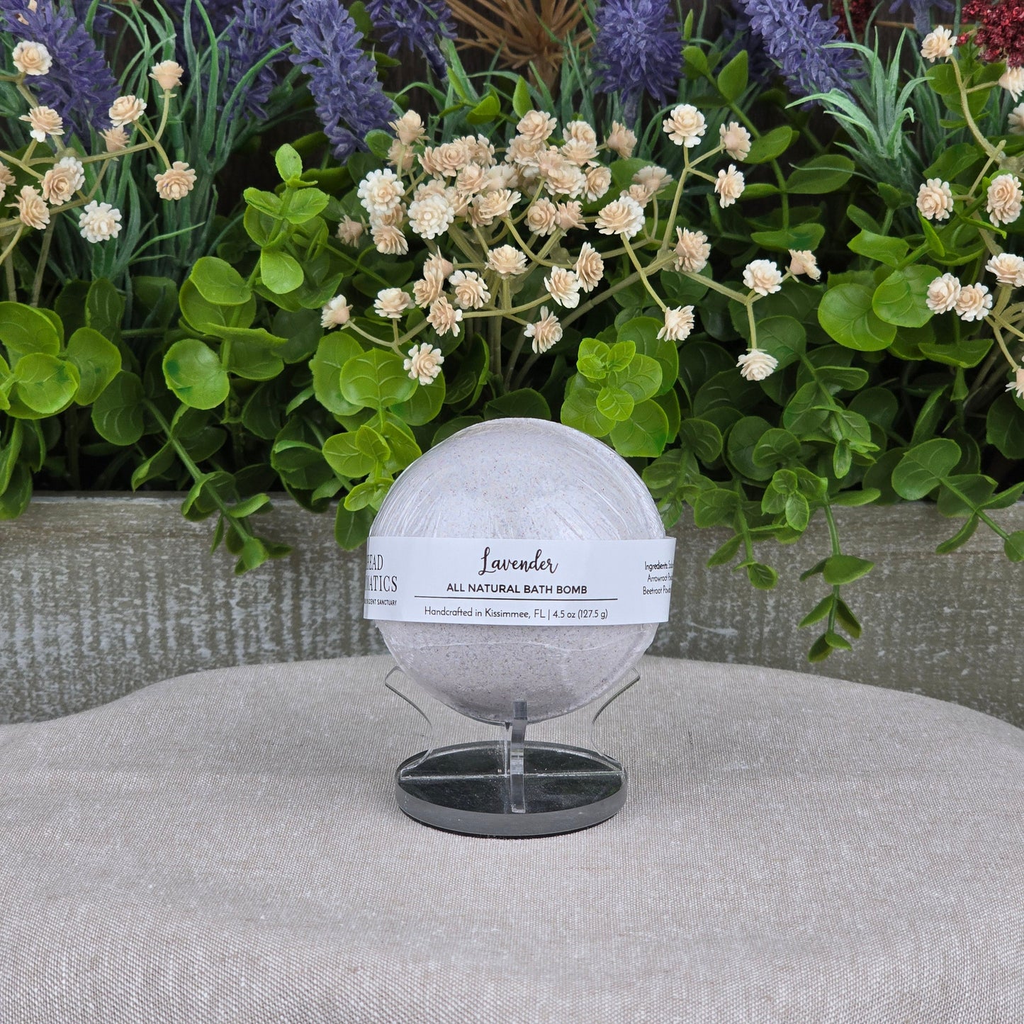 The Patchouli Bath Bomb - All Natural by Jarhead Aromatics rests on a black stand before lush artificial flowers and greenery, with a gray textured backdrop and beige fabric surface.