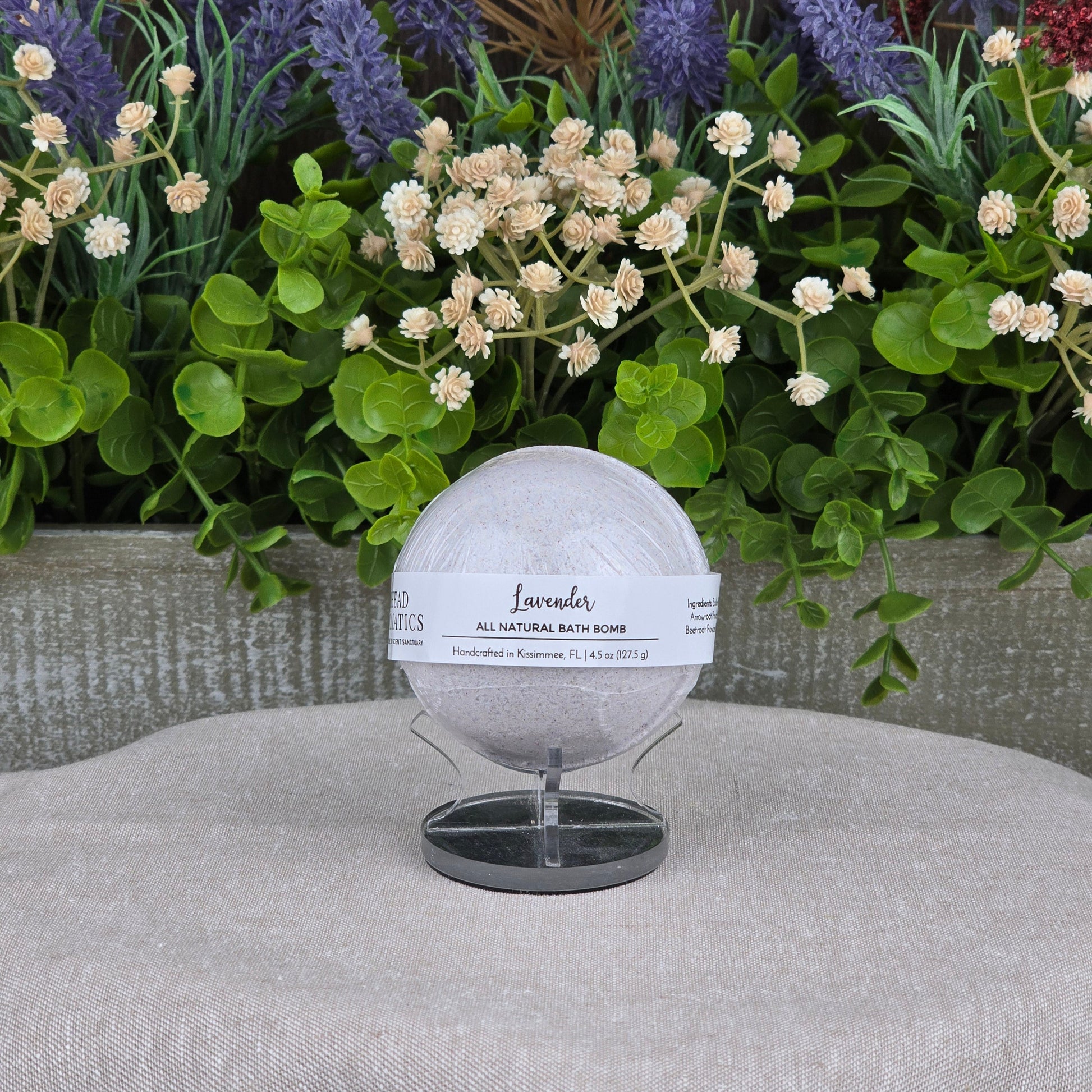 The Patchouli Bath Bomb - All Natural by Jarhead Aromatics rests on a black stand before lush artificial flowers and greenery, with a gray textured backdrop and beige fabric surface.