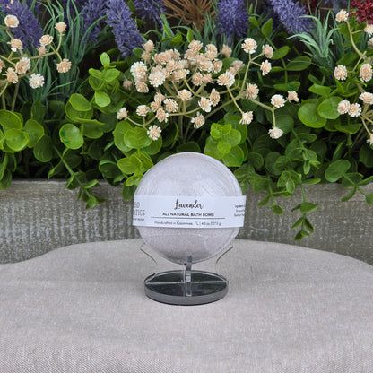 The Patchouli Bath Bomb - All Natural by Jarhead Aromatics rests on a black stand before lush artificial flowers and greenery, with a gray textured backdrop and beige fabric surface.