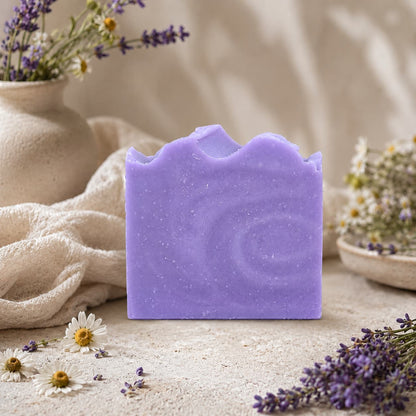 A bar of Jarhead Aromatics’ Lavender + Chamomile Handmade Coconut Milk Artisan Soap stands upright on a textured surface, surrounded by delicate wildflowers, lavender sprigs, and soft fabric, with beige pottery in the background. This calming herbal floral soap blends soothing botanicals for a gentle and luxurious bathing experience.
