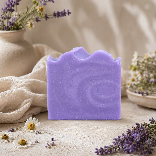 A bar of Jarhead Aromatics’ Lavender + Chamomile Handmade Coconut Milk Artisan Soap stands upright on a textured surface, surrounded by delicate wildflowers, lavender sprigs, and soft fabric, with beige pottery in the background. This calming herbal floral soap blends soothing botanicals for a gentle and luxurious bathing experience.