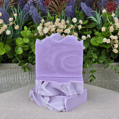 Two bars of Jarhead Aromatics Lavender + Chamomile Handmade Coconut Milk Artisan Soap with textured swirls are stacked on a beige surface amid green foliage and purple and white dried flowers.