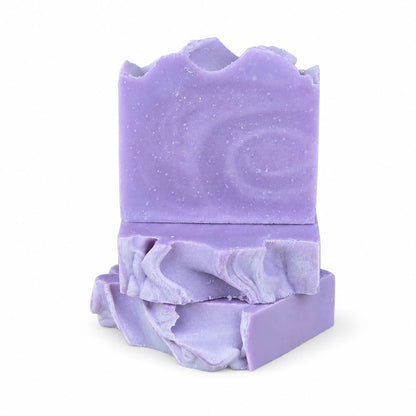 A bar of Jarhead Aromatics’ Lavender + Chamomile Handmade Coconut Milk Artisan Soap, tinted purple with a wavy textured top and two uneven broken pieces stacked below it, is displayed against a white background.