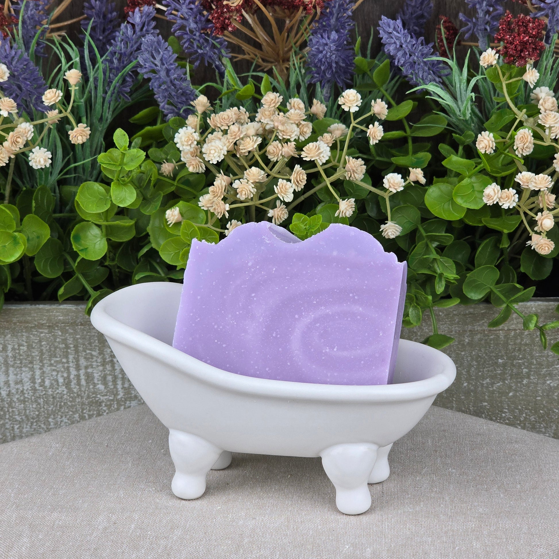 A bar of Jarhead Aromatics’ Lavender + Chamomile Handmade Coconut Milk Artisan Soap rests in a small white clawfoot bathtub dish, with green leaves and purple and white flowers in the background.