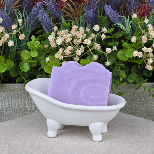 A bar of Jarhead Aromatics’ Lavender + Chamomile Handmade Coconut Milk Artisan Soap rests in a small white clawfoot bathtub dish, with green leaves and purple and white flowers in the background.