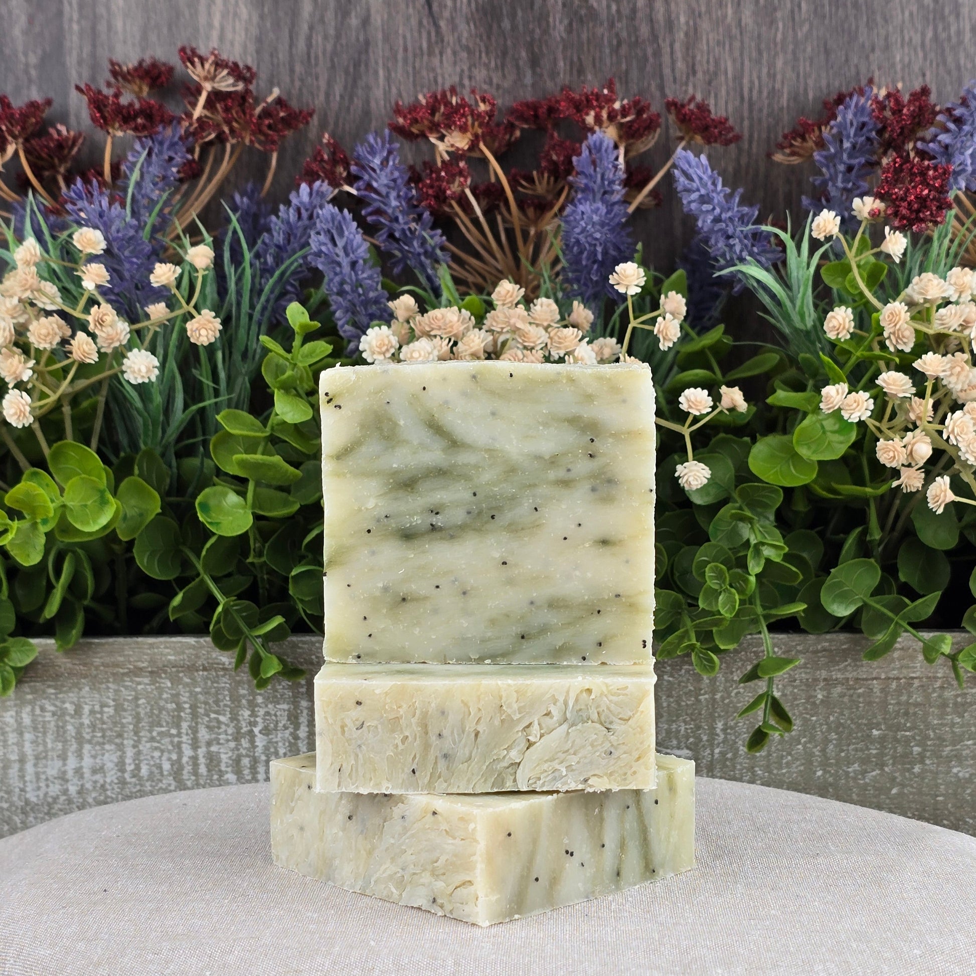 Three handmade Lavender + Eucalyptus soaps by Jarhead Aromatics, crafted with all-natural ingredients and featuring a marbled green and cream design, are stacked in front of artificial flowers on a light surface.