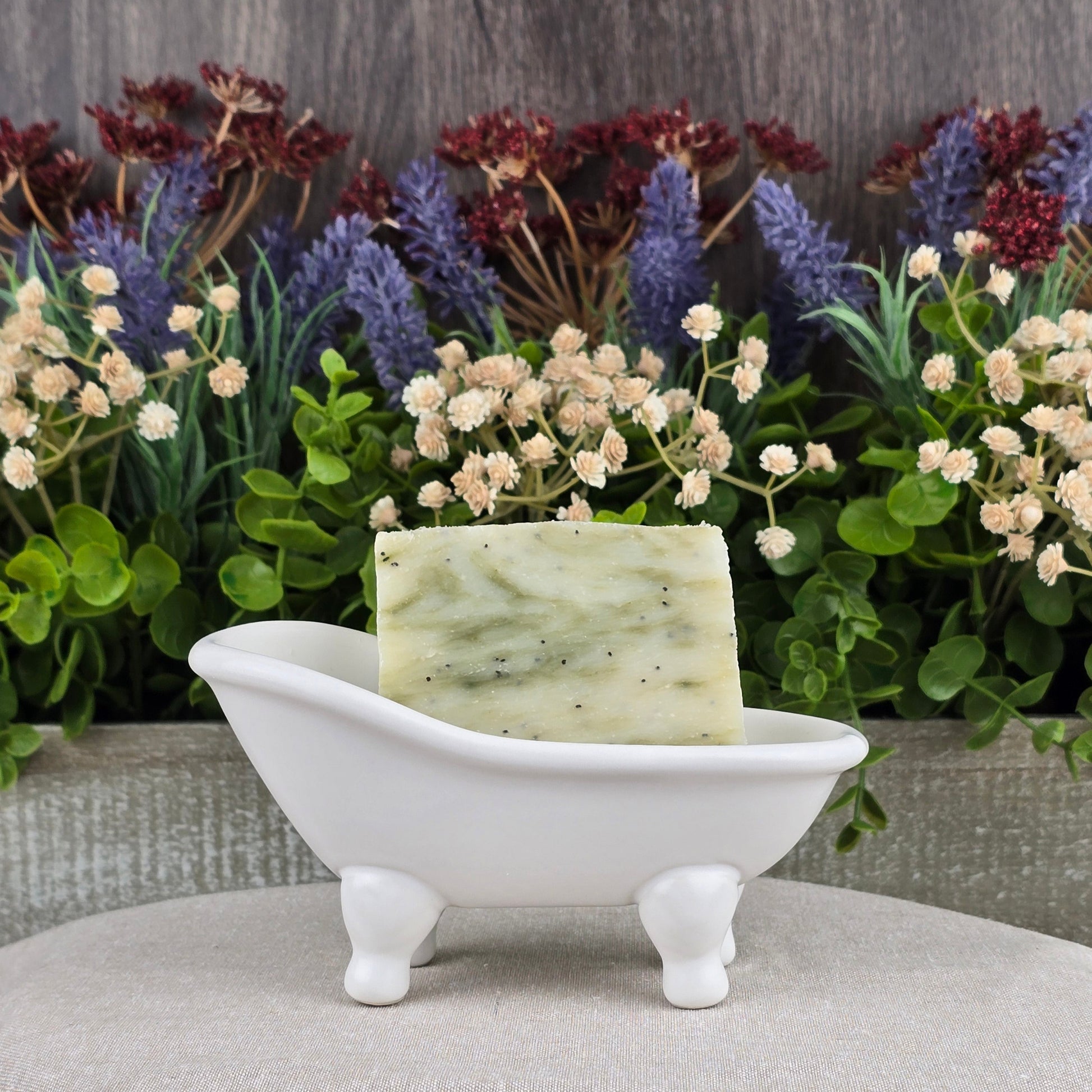 The Lavender + Eucalyptus Handmade Soap by Jarhead Aromatics rests in a white, miniature bathtub dish, surrounded by vibrant artificial flowers and lush green foliage.
