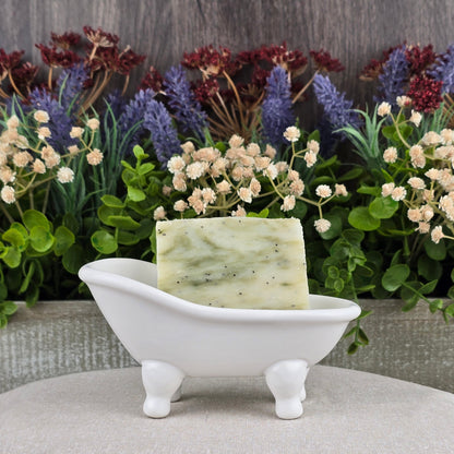 The Lavender + Eucalyptus Handmade Soap by Jarhead Aromatics rests in a white, miniature bathtub dish, surrounded by vibrant artificial flowers and lush green foliage.