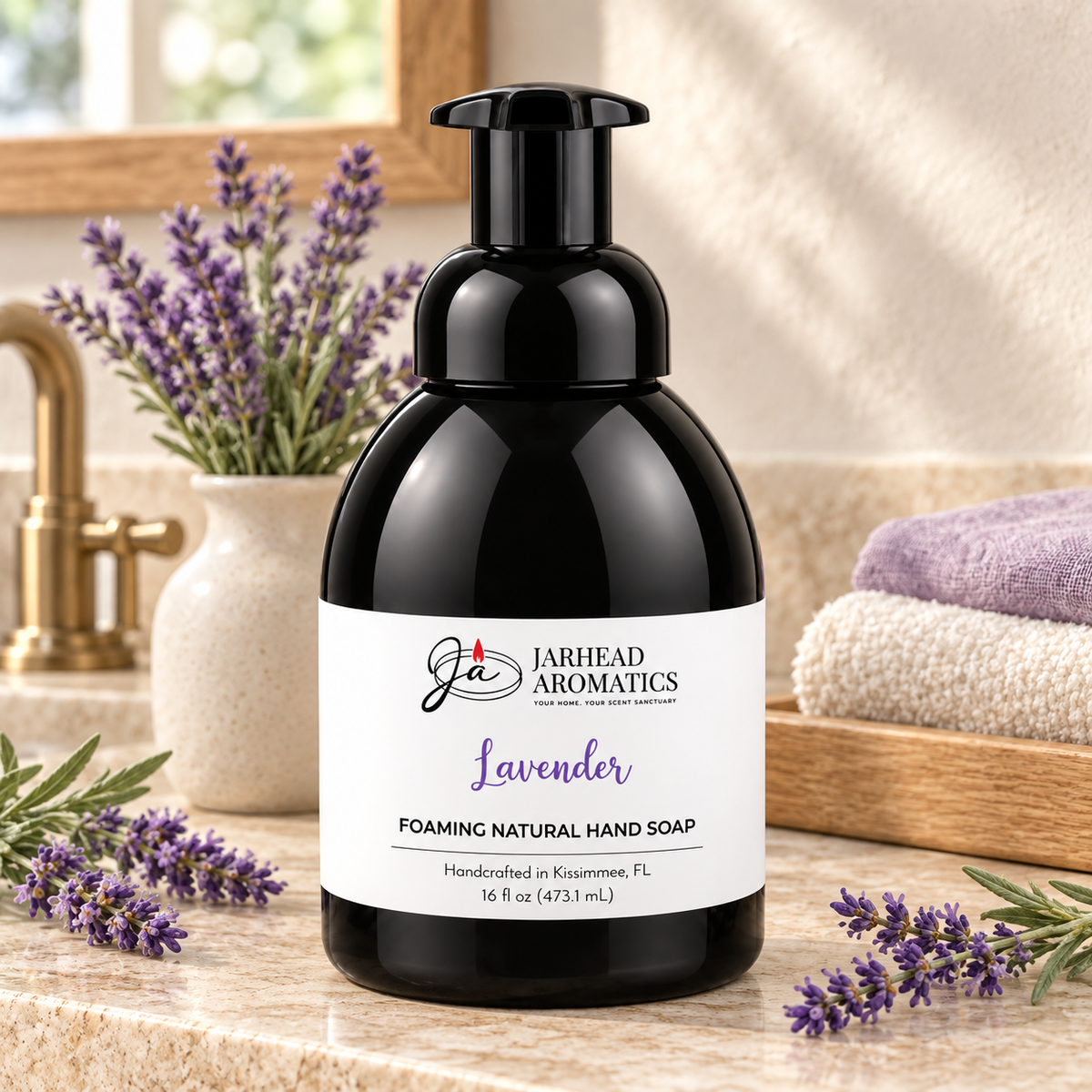 A black bottle of Lavender Handmade Foaming Natural Hand Soap by Jarhead Aromatics sits on a bathroom counter next to lavender sprigs and folded towels.