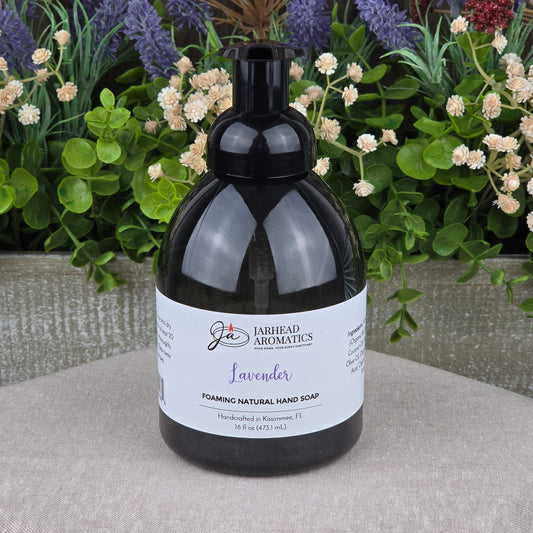 A bottle of Jarhead Aromatics Lavender Foaming Natural Hand Soap rests on a gray surface, with artificial lavender and colorful flowers in the background.