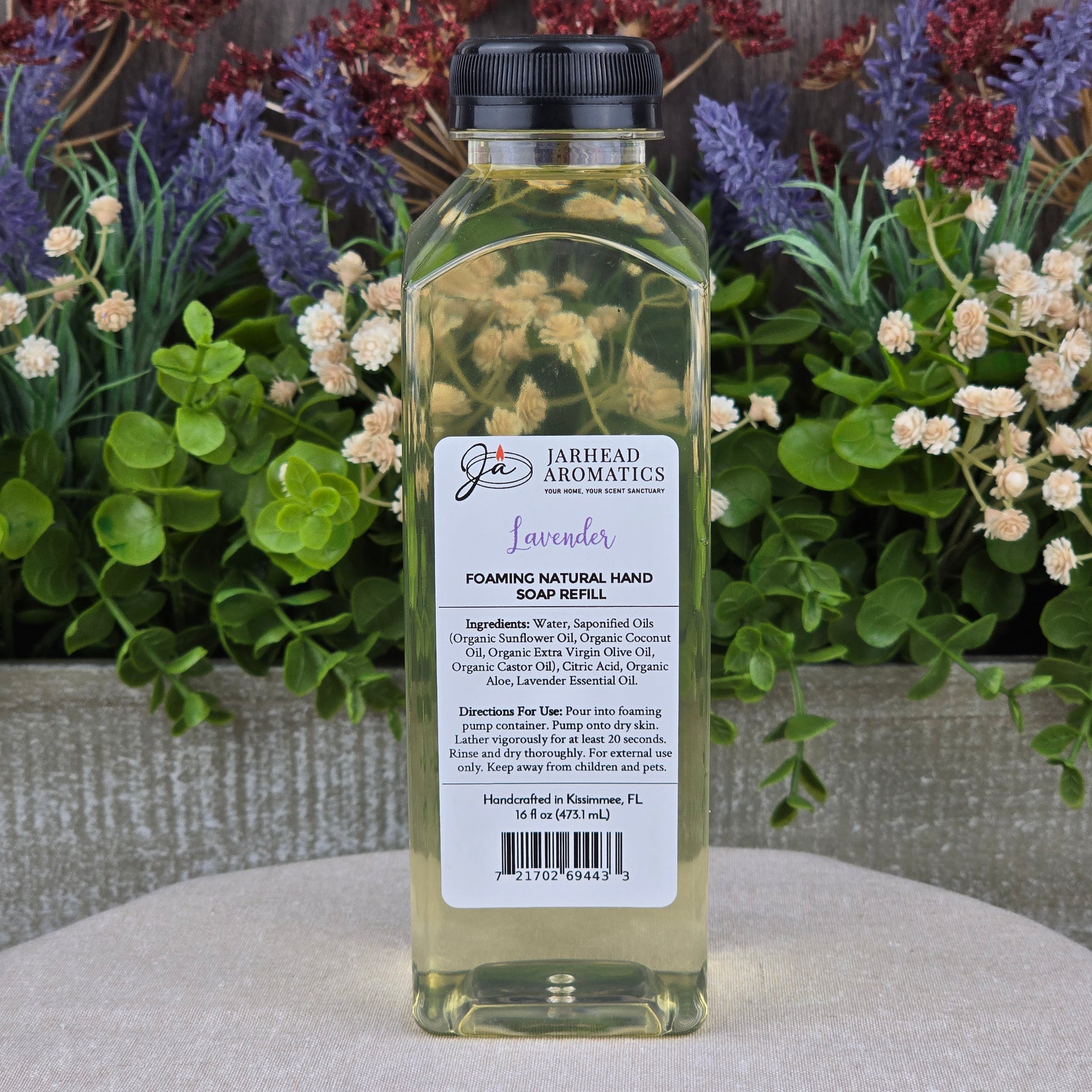 A clear plastic bottle of Jarhead Aromatics Lavender Foaming Natural Hand Soap sits on a gray surface, backed by purple and white flowers. The label notes its plant-based formula made with organic oils.
