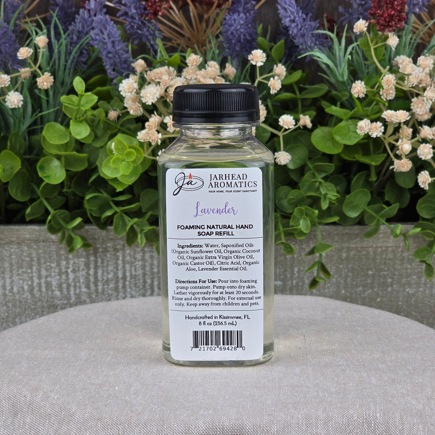 A clear bottle of Jarhead Aromatics Lavender Foaming Natural Hand Soap, made with plant-based ingredients, sits on a gray surface. Colorful artificial flowers and greenery form the backdrop, with the product label visible.