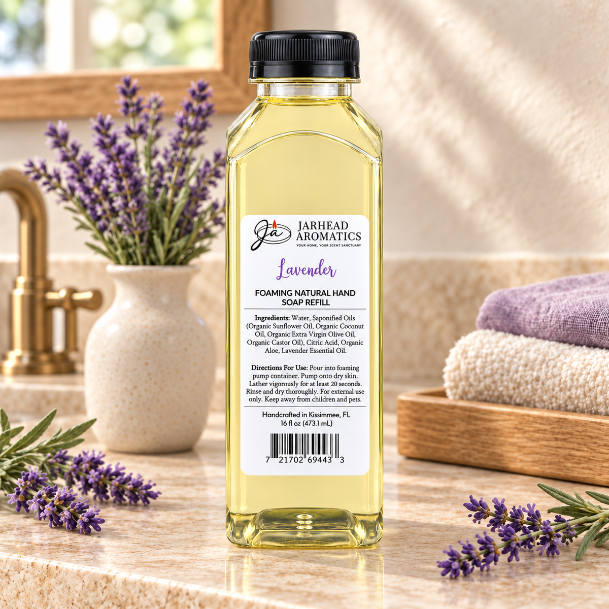 A clear plastic bottle of Jarhead Aromatics Lavender Handmade Foaming Natural Hand Soap sitting on a countertop next to lavender sprigs, a vase of lavender, and folded towels.