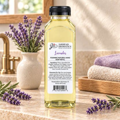 A clear plastic bottle of Jarhead Aromatics Lavender Handmade Foaming Natural Hand Soap sitting on a countertop next to lavender sprigs, a vase of lavender, and folded towels.