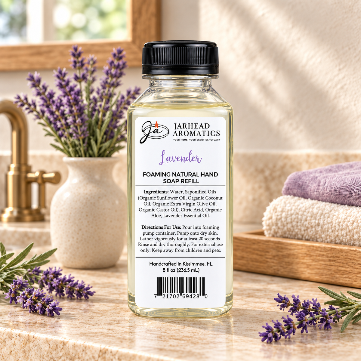 A clear bottle of Jarhead Aromatics Lavender Handmade Foaming Natural Hand Soap sits on a bathroom counter next to a vase of lavender sprigs and folded towels.