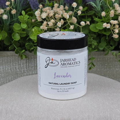 A 16 oz jar of Jarhead Aromatics Lavender Handmade Natural Laundry Soap sits on a beige surface with green and purple plants. This non-toxic detergent powder from Kissimmee, FL, cleans up to 32 loads.