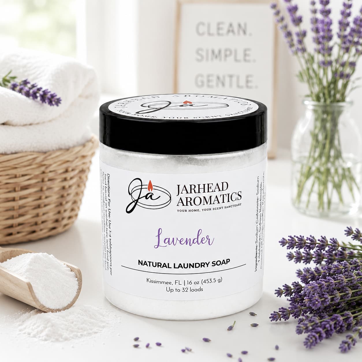 A jar of Jarhead Aromatics Lavender Handmade Natural Laundry Soap sits on a white table alongside a scoop of powder, fresh lavender sprigs, and a towel. In the background, a sign reads "Clean. Simple. Gentle." This powdered detergent with lavender essential oil provides a handmade and natural laundry solution for those who appreciate pure and simple ingredients.