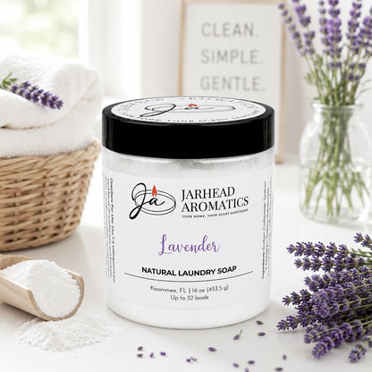 A jar of Jarhead Aromatics Lavender Handmade Natural Laundry Soap sits on a white table alongside a scoop of powder, fresh lavender sprigs, and a towel. In the background, a sign reads "Clean. Simple. Gentle." This powdered detergent with lavender essential oil provides a handmade and natural laundry solution for those who appreciate pure and simple ingredients.