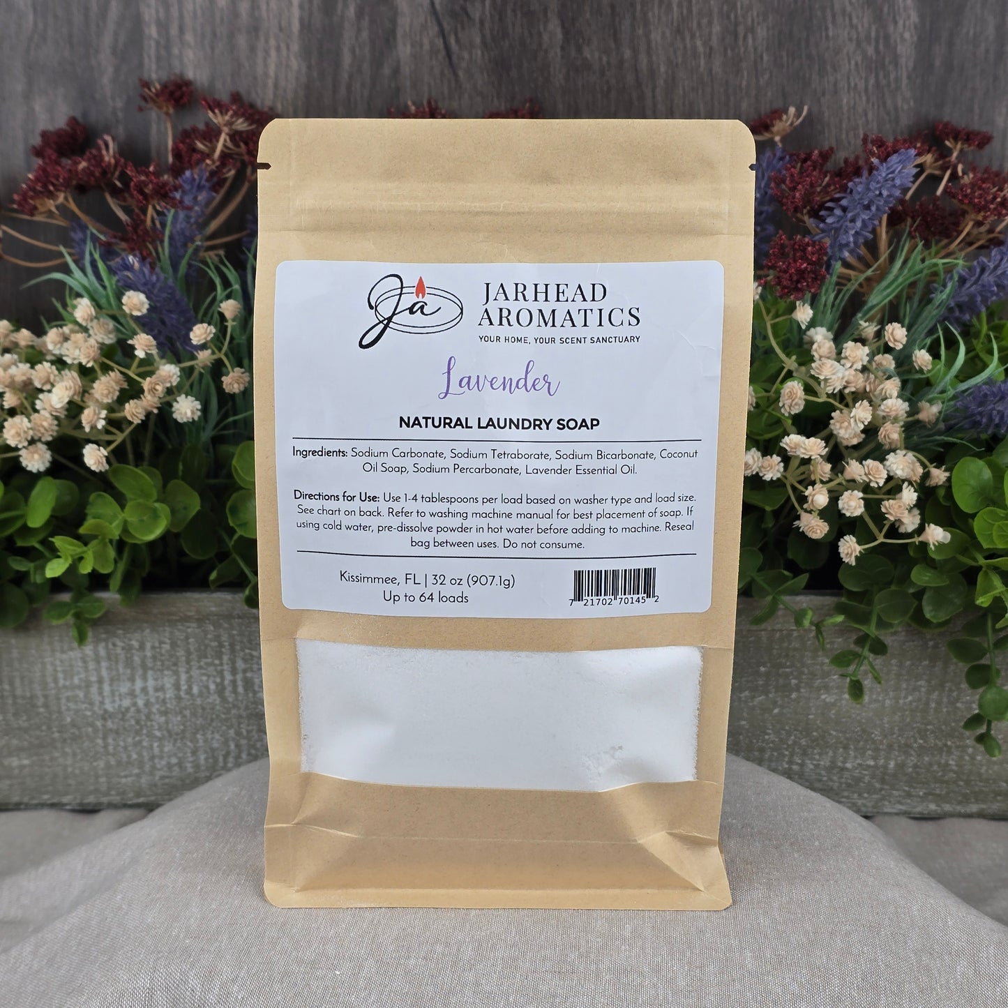 A brown paper pouch of Jarhead Aromatics Lavender Handmade Natural Laundry Soap is displayed upright among green foliage and purple flowers on a gray wood surface, with its label listing ingredients and usage instructions.