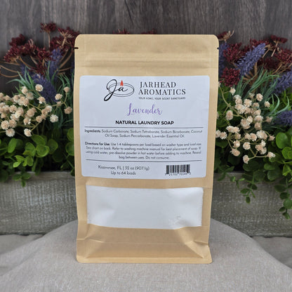 A brown paper pouch of Jarhead Aromatics Lavender Handmade Natural Laundry Soap is displayed upright among green foliage and purple flowers on a gray wood surface, with its label listing ingredients and usage instructions.