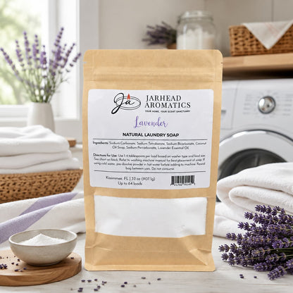 A bag of Jarhead Aromatics Lavender Handmade Natural Laundry Soap, a powdered detergent infused with lavender essential oil, rests on a table alongside fresh lavender sprigs, a towel, and a bowl of powder. The bright, clean setting highlights the product's label detailing ingredients and usage instructions.