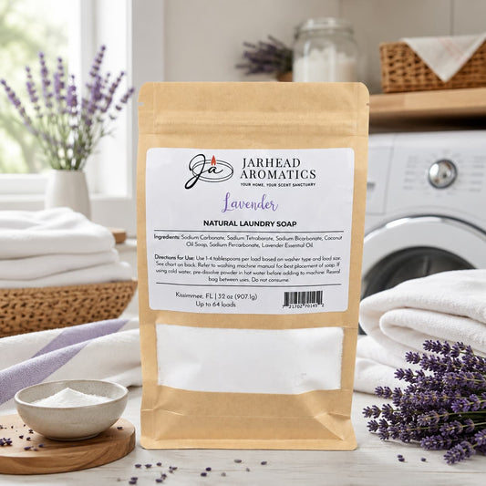 A bag of Jarhead Aromatics Lavender Handmade Natural Laundry Soap, a powdered detergent infused with lavender essential oil, rests on a table alongside fresh lavender sprigs, a towel, and a bowl of powder. The bright, clean setting highlights the product's label detailing ingredients and usage instructions.