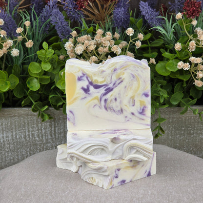 The Lavender + Lemon Handmade Coconut Milk Artisan Soap by Jarhead Aromatics features purple and yellow swirls on a creamy base, showcased with lush greenery and dried flowers atop a neutral fabric.