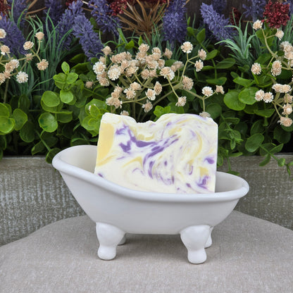 A bar of Jarhead Aromatics' Lavender + Lemon Handmade Coconut Milk Artisan Soap rests in a white ceramic bathtub, accented by green plants and purple flowers in the background.