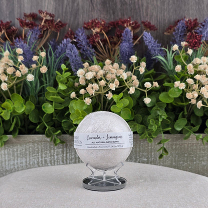 The Jarhead Aromatics Lavender + Lemongrass Bath Bomb - All Natural rests on a stand atop a beige cushion, with artificial flowers and greenery behind it to emphasize its natural ingredients.
