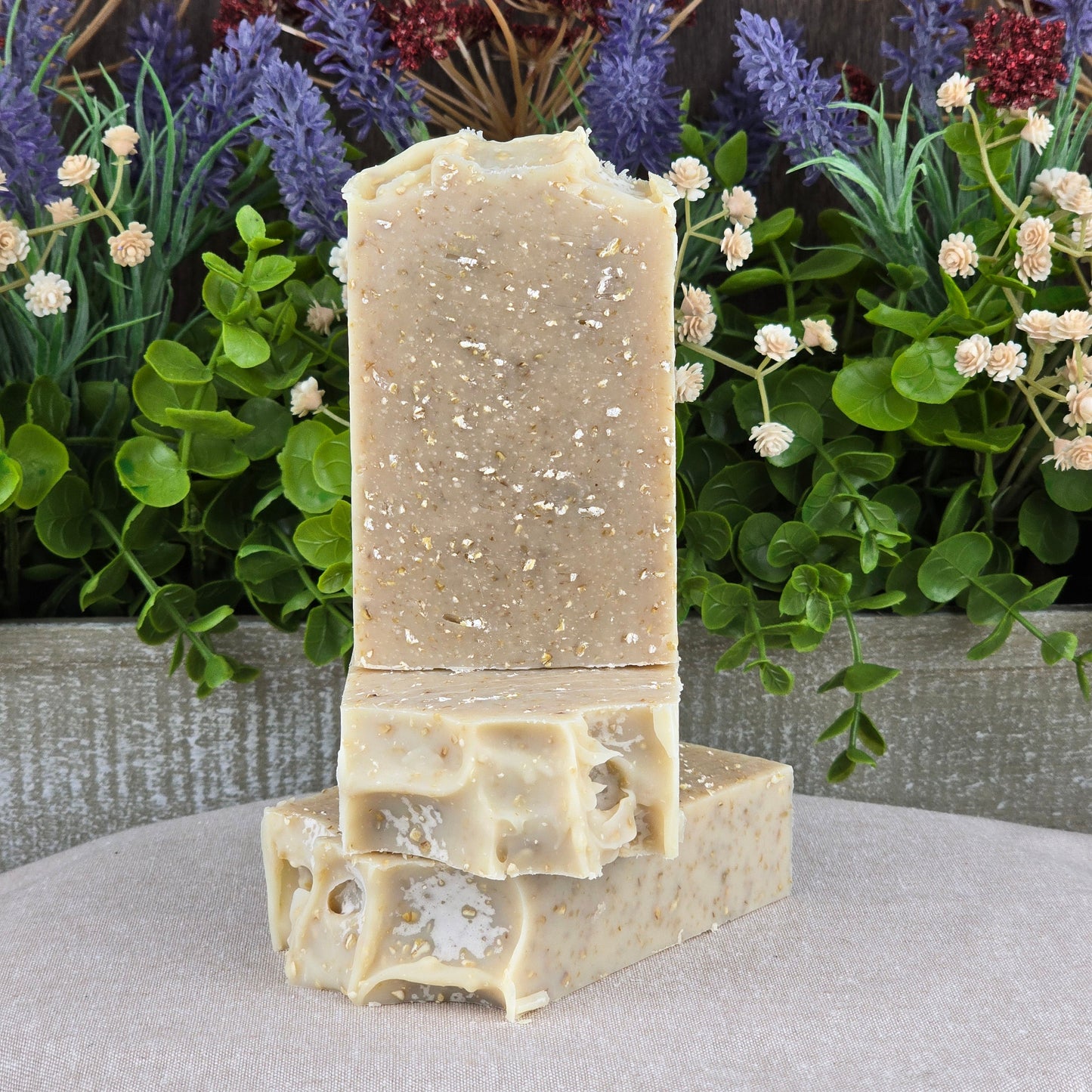 Two rectangular bars of Jarhead Aromatics Lavender + Oatmeal Handmade Artisan Soap – All Natural are stacked with lush green plants and purple flowers behind them. The speckled, natural beige soap reflects its soothing lavender essence.