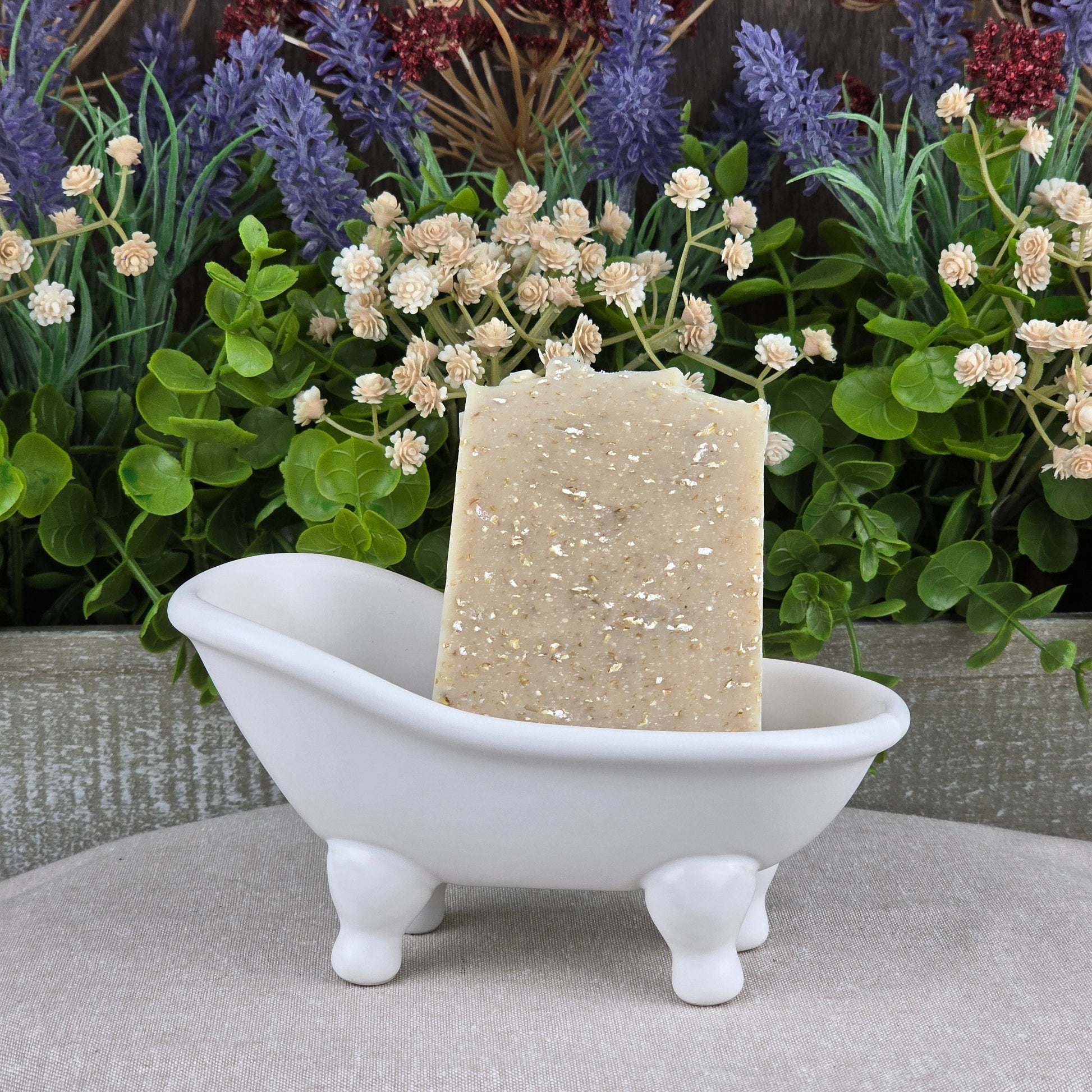 A bar of Jarhead Aromatics Lavender + Oatmeal Handmade Artisan Soap stands upright in a small white clawfoot bathtub soap dish, with artificial green plants and purple and white flowers in the background.