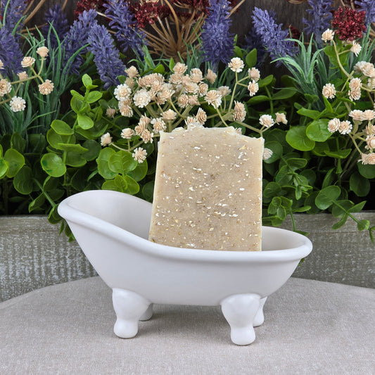 A bar of Jarhead Aromatics Lavender + Oatmeal Handmade Artisan Soap stands upright in a small white clawfoot bathtub soap dish, with artificial green plants and purple and white flowers in the background.
