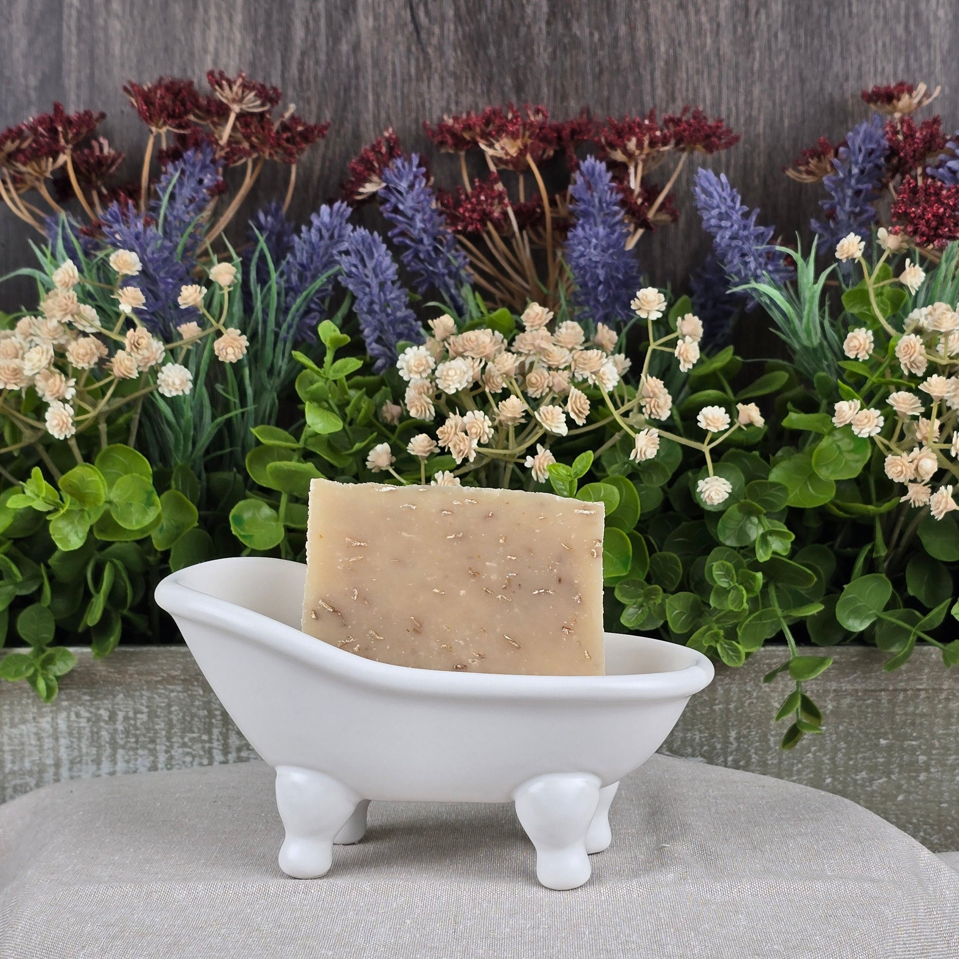 The Lavender + Oatmeal Handmade Goat Milk Soap by Jarhead Aromatics sits in a decorative white bathtub on a textured surface, complemented by vibrant artificial lavender and red-tipped foliage, adding rustic charm against a wooden backdrop.