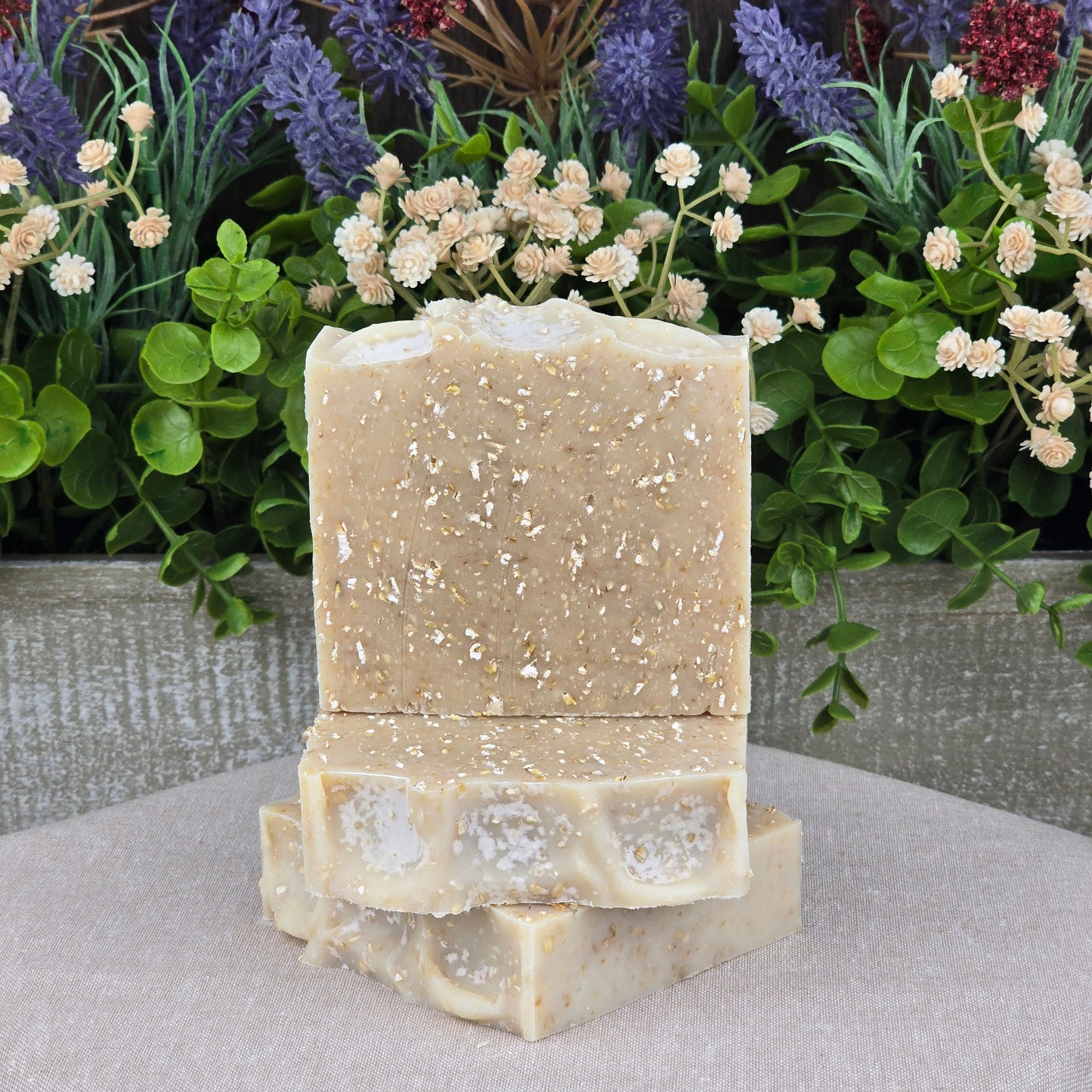 Three handmade Lavender + Oatmeal Natural Goat Milk Artisan Soaps by Jarhead Aromatics, featuring visible oat flakes, are stacked on a neutral cloth in front of green foliage and colorful artificial flowers.