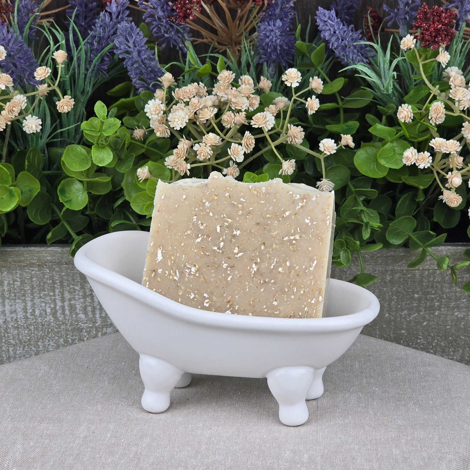 A bar of Jarhead Aromatics Lavender + Oatmeal Handmade Natural Goat Milk Artisan Soap sits in a small white clawfoot bathtub dish, surrounded by artificial flowers and greenery in the background.