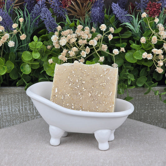 A bar of Jarhead Aromatics Lavender + Oatmeal Handmade Natural Goat Milk Artisan Soap sits in a small white clawfoot bathtub dish, surrounded by artificial flowers and greenery in the background.