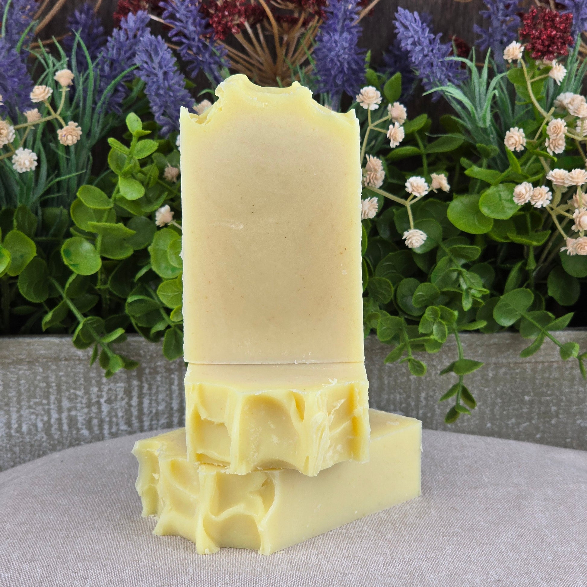 Three Lavender + Peppermint Handmade Artisan Soap bars from Jarhead Aromatics are stacked in front of lush green leaves and purple and white flowers, with one all-natural soap bar standing upright on top.