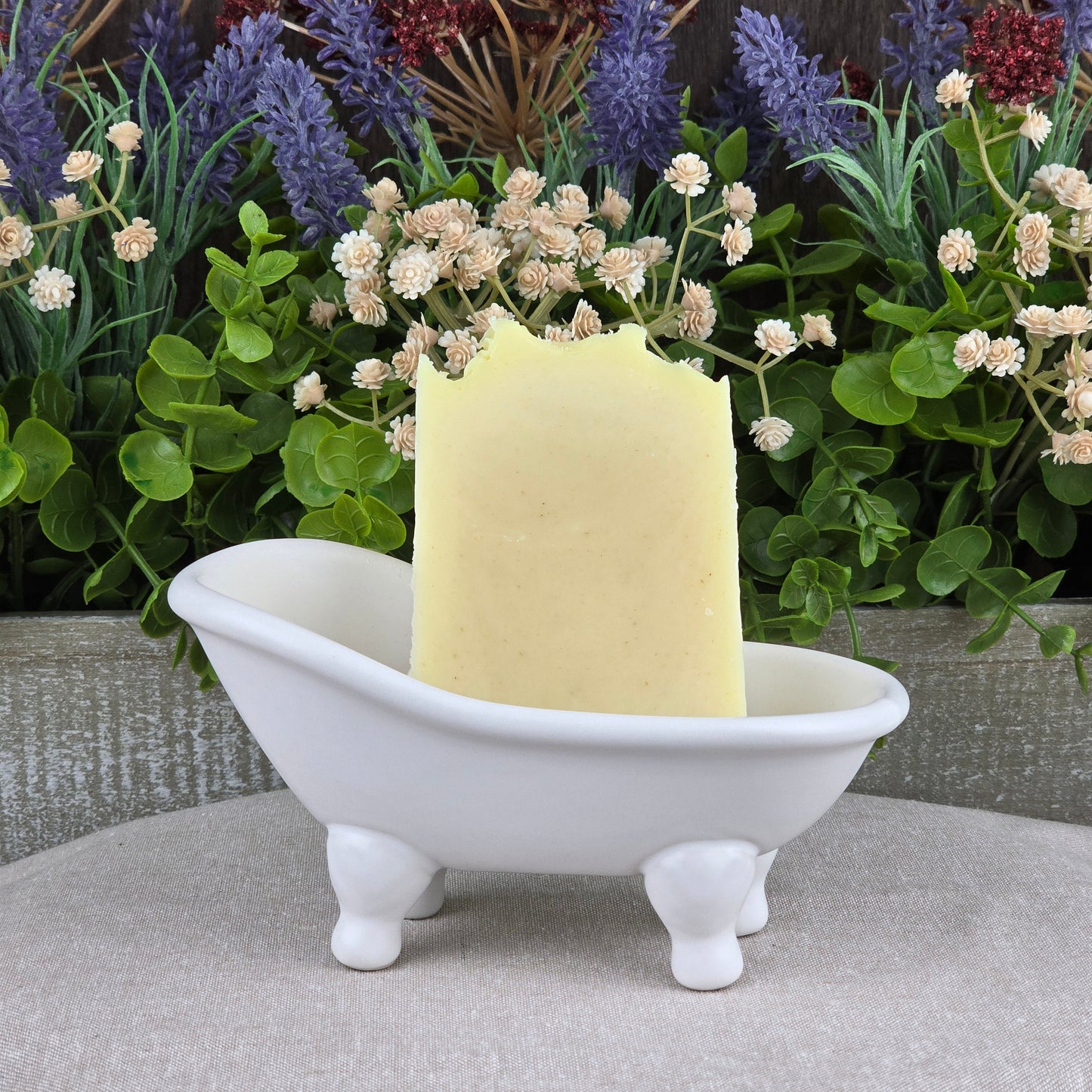 A rectangular bar of Jarhead Aromatics' Lavender + Peppermint Handmade Artisan Soap stands upright in a small white clawfoot bathtub-shaped dish, with lush green and purple artificial flowers arranged in the background.