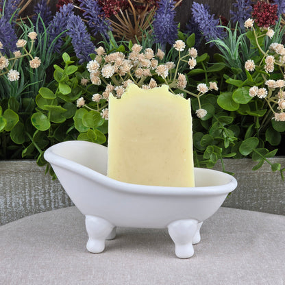A rectangular bar of Jarhead Aromatics' Lavender + Peppermint Handmade Artisan Soap stands upright in a small white clawfoot bathtub-shaped dish, with lush green and purple artificial flowers arranged in the background.