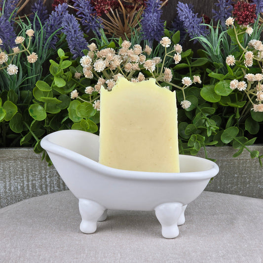 A rectangular bar of Jarhead Aromatics' Lavender + Peppermint Handmade Artisan Soap stands upright in a small white clawfoot bathtub-shaped dish, with lush green and purple artificial flowers arranged in the background.
