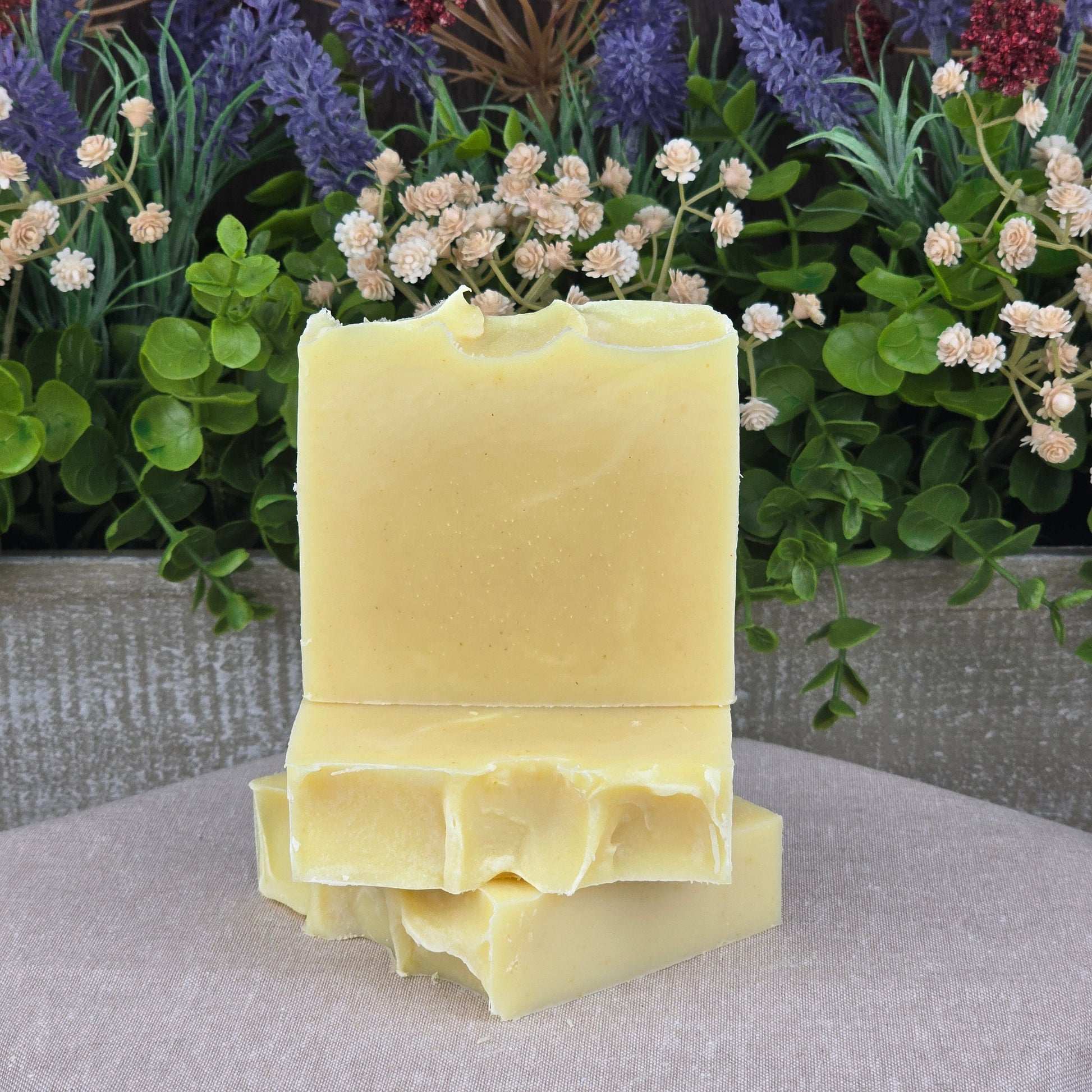 Three bars of Jarhead Aromatics’ Lavender + Peppermint Handmade Natural Artisan Soap are stacked on beige cloth with green leaves and purple, red, and white dried flowers—highlighting their handcrafted quality.